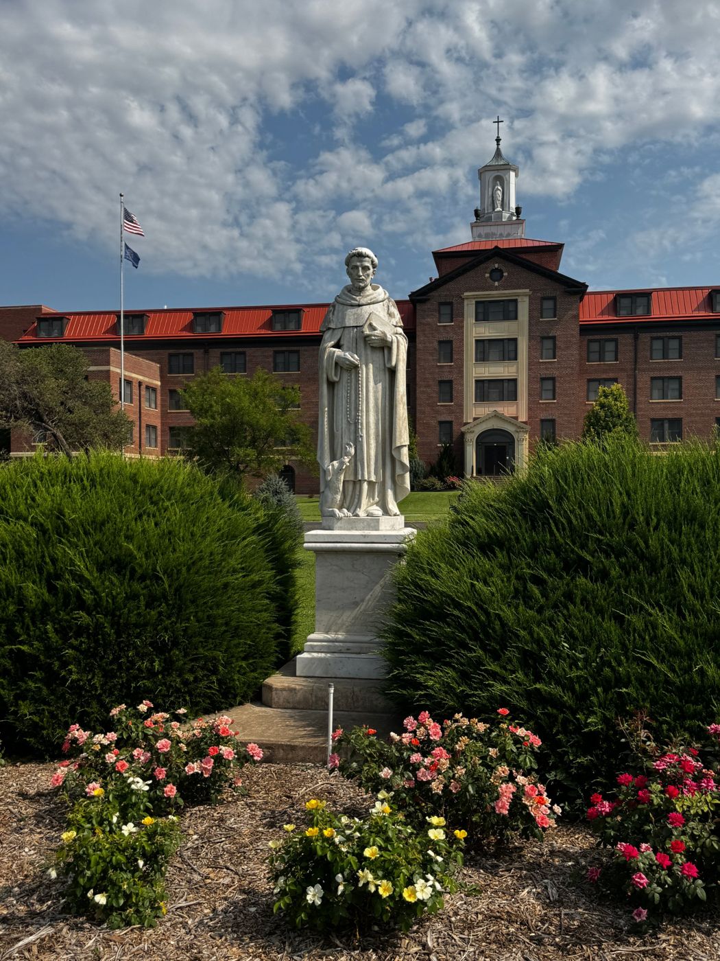 The red-brick building at 3600 Broadway with its white cupola, seen across the front garden with a marble statue and rose beds